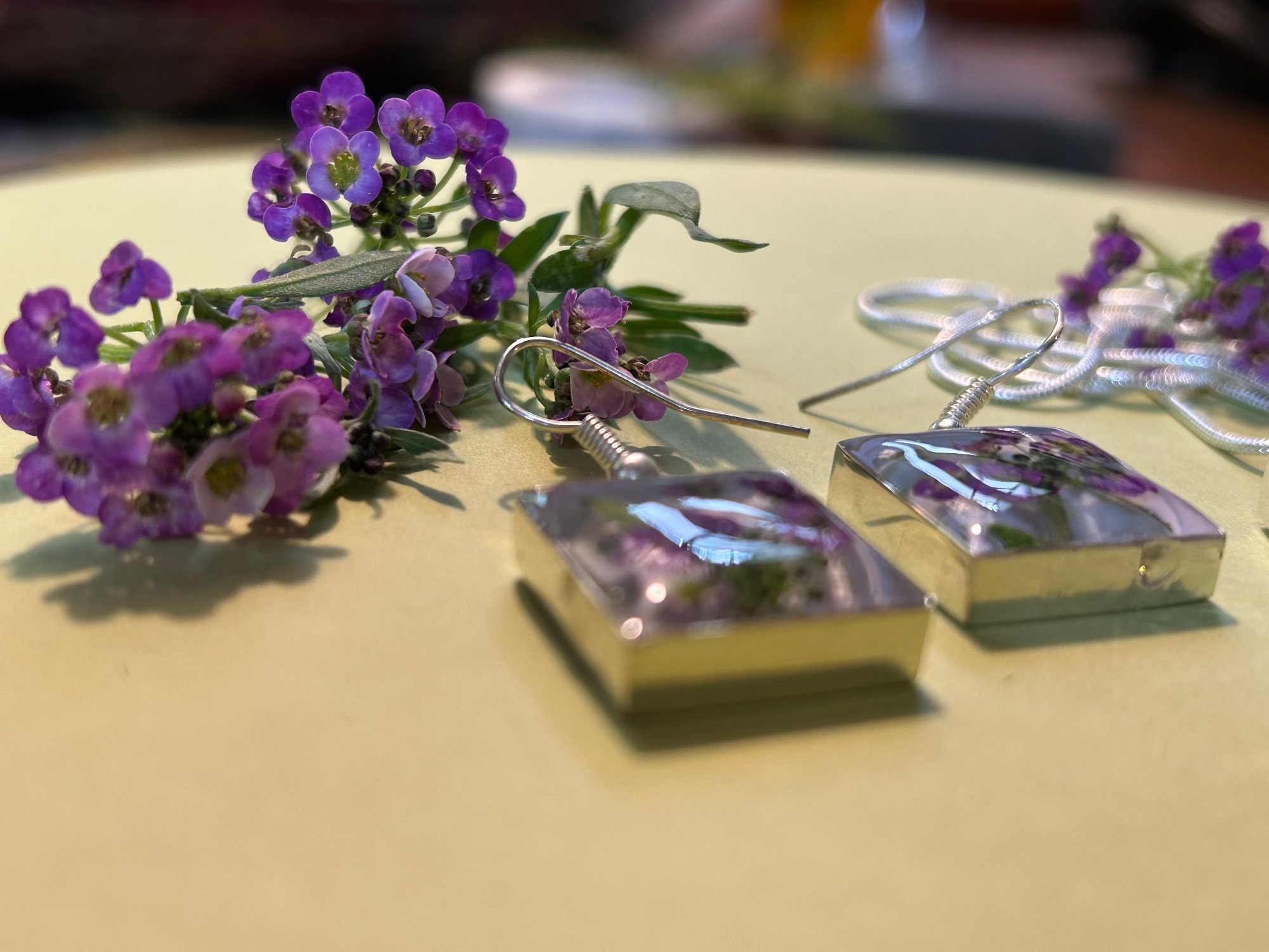 Silver earrings with embedded flowers on a beige surface with purple flowers.